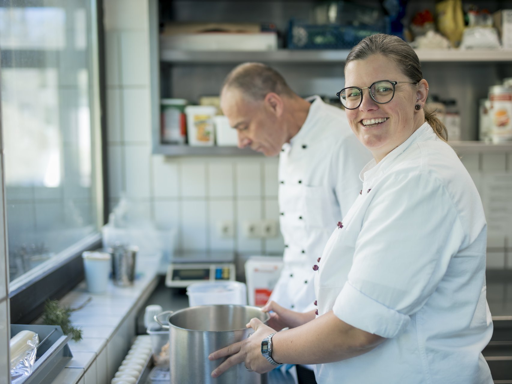 Eine Frau lächelt in die Kamera, während sie mit einem Mann in der Küche arbeitet.
