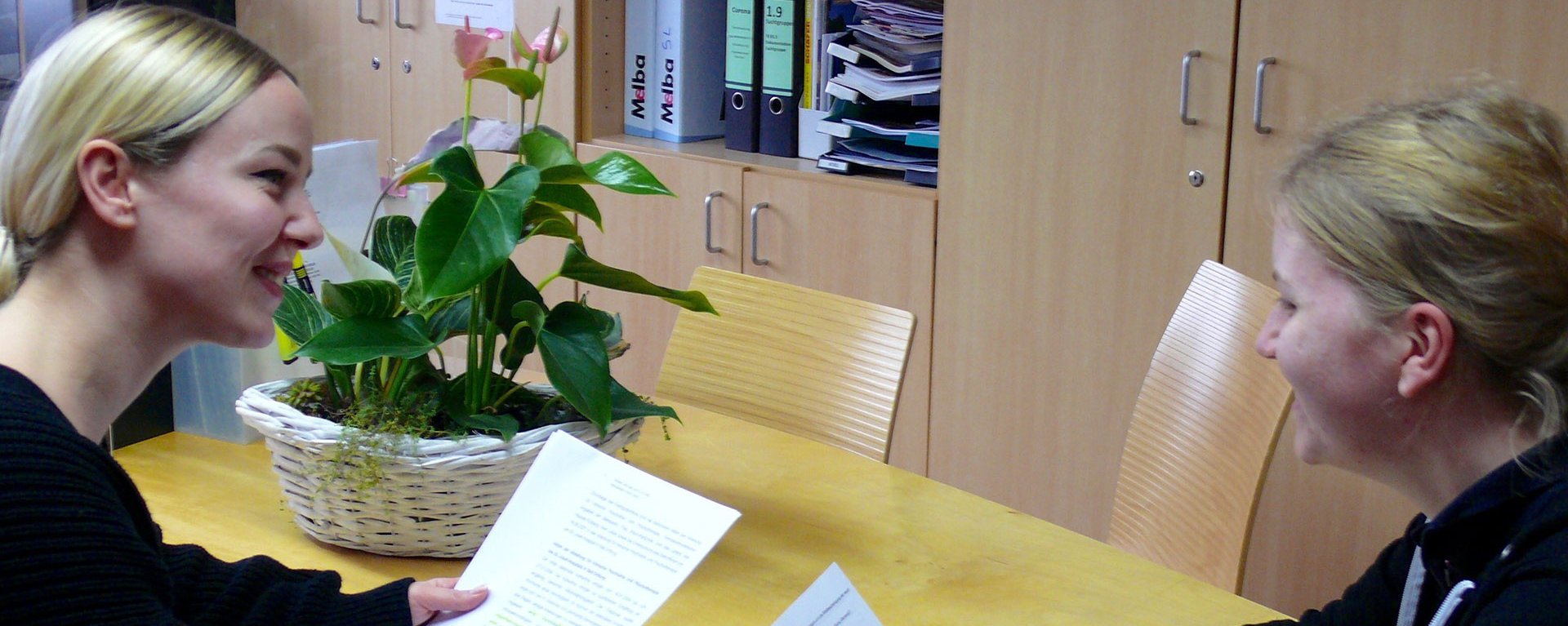 Im Büro: zwei Frauen halten jeweils ein Blatt Papier und unterhalten sich