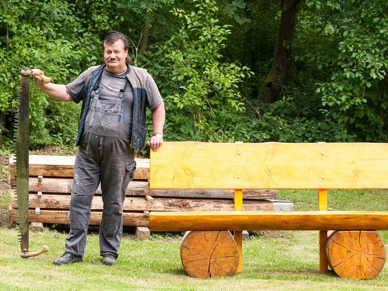 ein Mann in Arbeitskleidung lehnt an einer Holzbank - in der freien Hand eine mannshohe Baumsäge